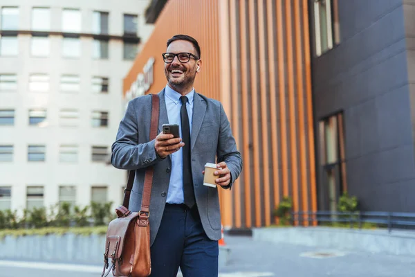 Takım elbiseli neşeli bir işadamı elinde kahve fincanı ve akıllı telefonuyla modern bir ofis binasının önünde yürüyor. Kendine güveni ve başarısı var, işte verimli bir günün tadını çıkarıyor..