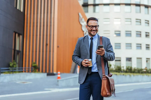 Gri takım elbiseli kendine güvenen bir işadamı akıllı telefonunu kullanıp elinde kahve tutarken yürüyor. Kentsel ortam profesyonel atmosferi geliştiriyor, dinamik bir sabah rutini sergiliyor..