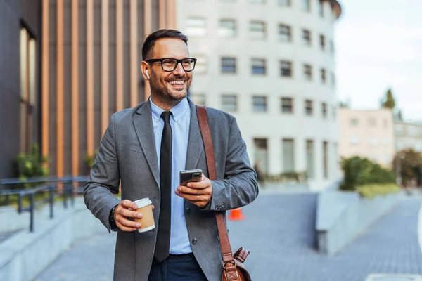 Gülümseyen bir iş adamı elinde kahve fincanı ve akıllı telefonuyla dışarı çıkar. Takım elbise giymiş, kendinden emin ve bağlı görünüyor, kablosuz kulaklıklarla, modern ofis arkaplanına karşı..