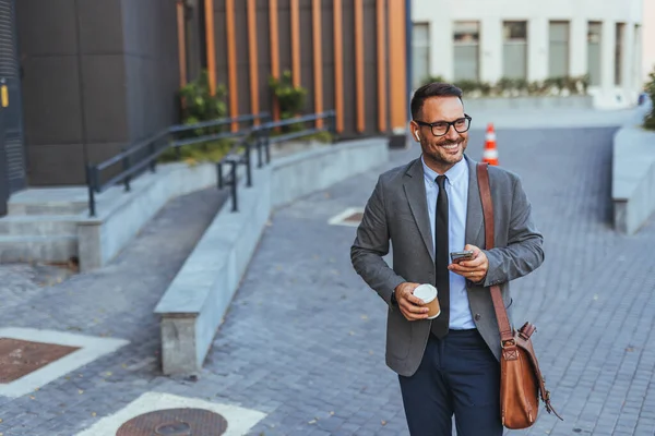 Resmi giyinmiş neşeli bir işadamı, aydınlık bir günde güven ve üretkenlik saçarak şehir ortamında yürürken bir kahvenin tadını çıkarır ve akıllı telefonunu kullanır..