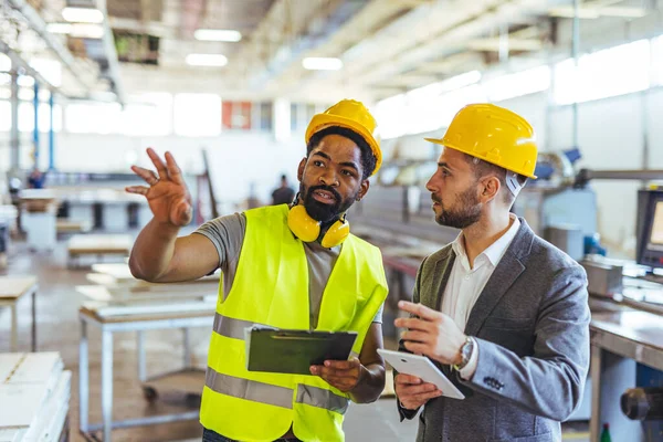 İki profesyonel, büyük bir sanayi tesisindeki operasyonlar hakkında sohbet ediyorlar. Biri pratik ve güvenliği sembolize eden bir güvenlik yeleği giyerken, diğeri takım çalışması ve teknik uzmanlığı vurgulayarak dikkatle iş birliği yapıyor..