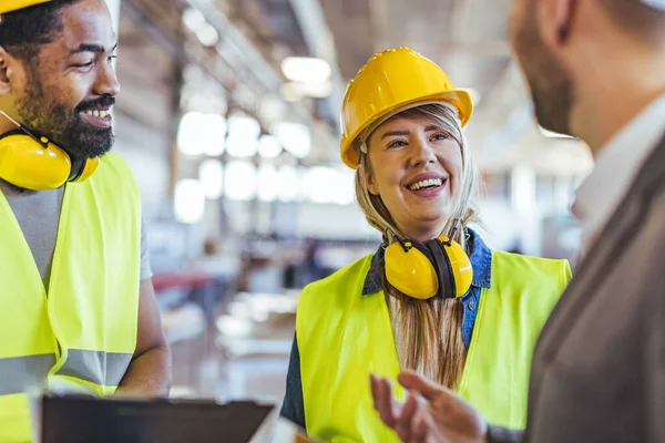Güvenlik teçhizatı takan ve bir depo içinde dostça bir sohbete girişen bir endüstri işçisi ekibi profesyonel bir ortamda takım çalışmasını ve işbirliğini teşvik ediyor..