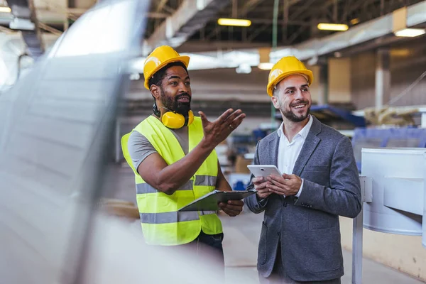 Güvenlik kaskı ve yelek giyen iki profesyonel, dijital tablet ve not panosuyla bir fabrikada işbirliği yaparak bina planlarına odaklanıyor. İnşaat ve imalatta takım çalışmasını, üretkenliği ve yeniliği sembolize ederler..
