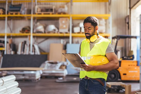 Depoda çalışan bir işçi, yansıtıcı bir güvenlik yeleği giyerken ve kask tutarken bir panoyu incelerken endüstrinin profesyonelliğini ve iş güvenliğine bağlılığını gösteriyor..
