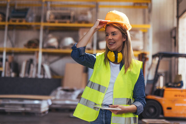 A cheerful construction worker wearing a hard hat and reflective vest stands in a warehouse. She exudes confidence and professionalism, symbolizing safety, skill, and teamwork in an industrial environment.