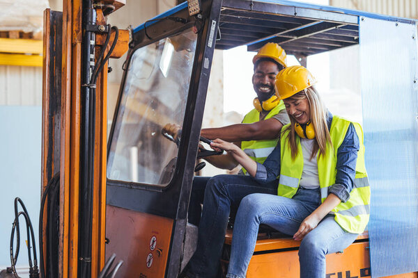 Two factory workers in safety equipment share a joyful moment while operating a forklift in the warehouse.