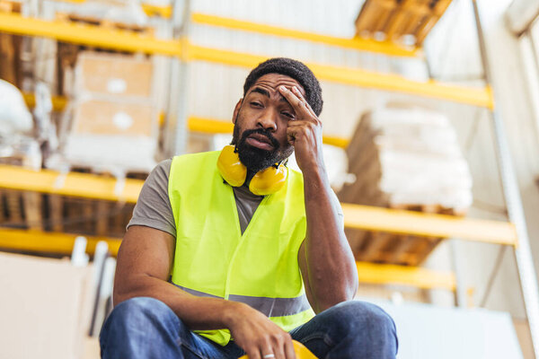A man wearing a high-visibility vest and safety headphones in a warehouse setting expresses stress, fatigue, or frustration.