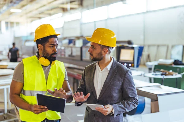 Güvenlik donanımı giyen iki profesyonel endüstriyel üretim binasında bir projeyi tartışıyor..