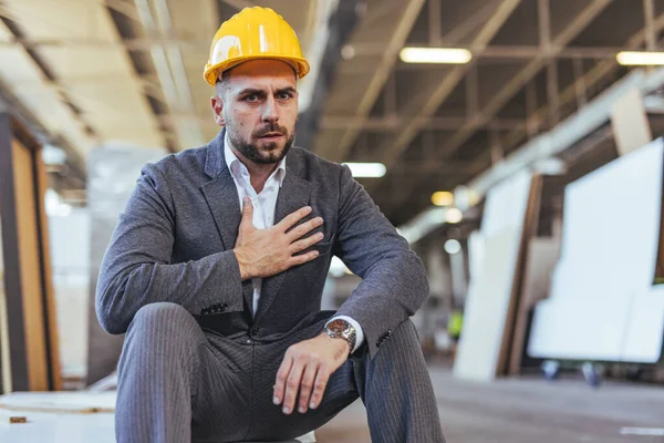 Güvenlik kaskıyla resmi iş kıyafeti giyen bir inşaat işçisi, kapalı bir endüstriyel iş sahasında endişe ve stresi dile getiriyor..