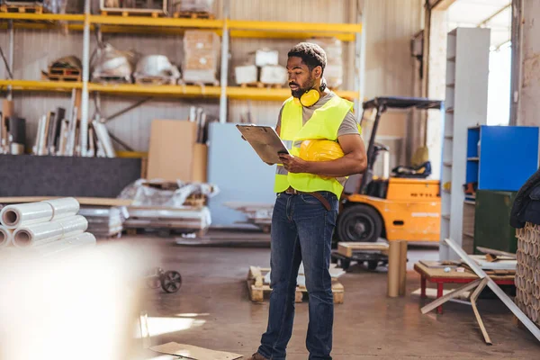 Envanterle çevrili endüstriyel bir depodaki panoyu inceleyen yansıtıcı yelek giyen bir işçi. Sahne, profesyonel bir çalışma ortamında verimliliği ve dikkati detaylara taşır.