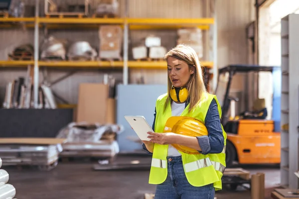 Güvenlik yeleği giyen ve organize bir depoda tablet kullanan bir kadın işçi. Kurulumda, sanayi ve lojistik ortamların vurgulandığı kutuları ve forklifti olan bir raf sistemi de yer alıyor..
