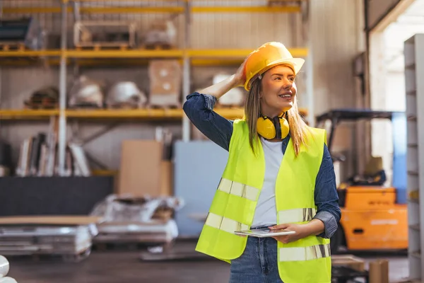 Bir depoda çalışan neşeli bir bayan işçi, yansıtıcı giysiler ve sıkı bir şapka giyiyor, endüstriyel bir ortamda güvenle ayakta duruyor. Güvenlik, verimlilik ve işte olumlu bir tavır..