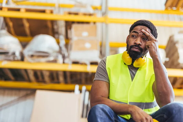 Depoda oturan yorgun bir işçi, yüksek görüş açısına sahip bir yelek ve düşünceli bir ifadeyle koruyucu giysiler giyiyor, raflarla çevrili, sıkı çalışmayı, yorgunluğu ve kararlılığı simgeliyor..