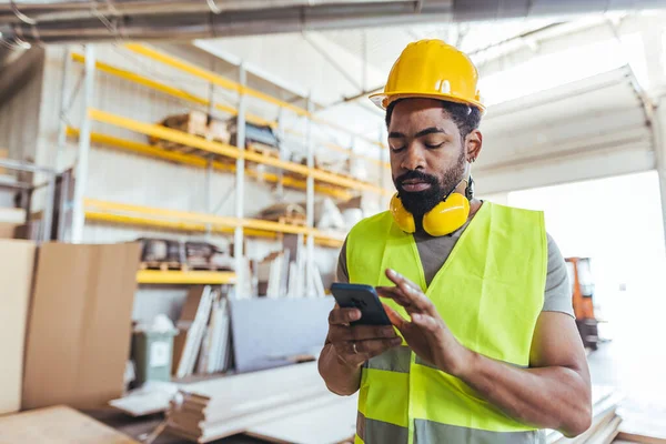 Sert şapka ve yansıtıcı yelek giyen bir işçi depoda akıllı telefon kullanıyor..