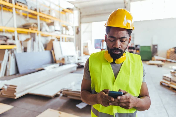 Yüksek görüşlü güvenlik elbiseleri ve miğferi olan bir inşaat işçisi depoda akıllı telefon kullanır. Görüntü sanayi ortamında profesyonellik, teknoloji ve işyeri güvenliğini yansıtıyor.