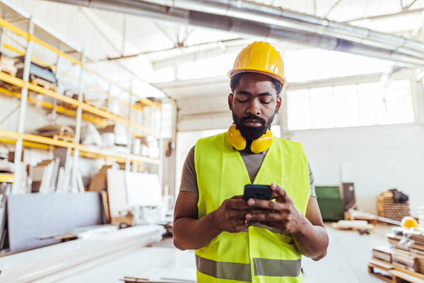 A construction worker wearing safety gear and using a smartphone inside a well-lit workshop. Various work materials are visible in the background, creating an industrial and professional atmosphere.