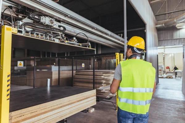 Güvenlik teçhizatı takan bir işçi endüstriyel bir ortamda, üretim ve işyeri güvenliğini vurgulayarak, makineleri izler. Tesiste modern ekipman ve organize operasyonlar imalat ortamında sergileniyor..