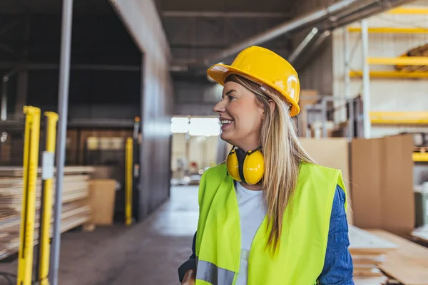 Üretim tesisinde gülümseyen, iş yerinde olumlu ve profesyonellik gösteren, sağlam bir şapka takan, yüksek görüşlü bir yelek giyen kendine güvenen bir kadın..