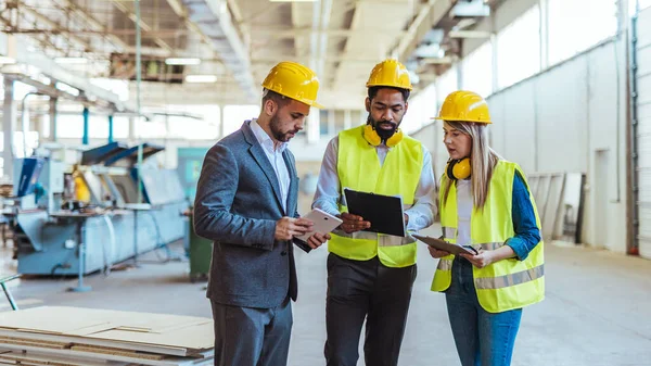 Çeşitli fabrika çalışanlarından oluşan bir ekip ve bir üretim tesisi içinde takım çalışması, liderlik ve endüstriyel güvenliği temsil eden, kask ve yüksek görünürlüklü yelek gibi koruyucu ekipmanlar giyen bir yönetici..