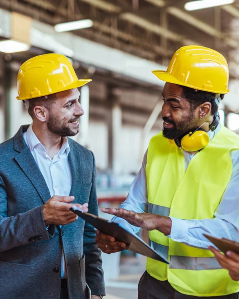 Gülümseyen Afrikalı Amerikalı erkek mimar ve miğfer ve güvenlik yeleği giymiş mühendis fabrikada çalışırken iş arkadaşıyla konuşuyor.