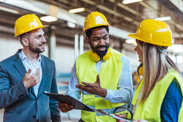 Çeşitli fabrika çalışanlarından oluşan bir ekip ve bir üretim tesisi içinde takım çalışması, liderlik ve endüstriyel güvenliği temsil eden, kask ve yüksek görünürlüklü yelek gibi koruyucu ekipmanlar giyen bir yönetici..