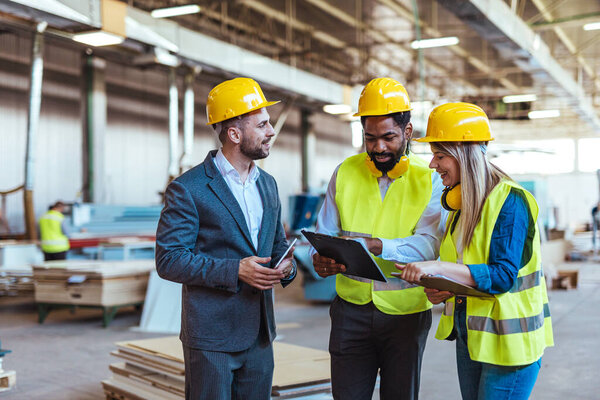 Diverse team of factory employees and a manager inside a production facility, representing teamwork, leadership, and industrial safety, wearing protective equipment like helmets and high-visibility vests.