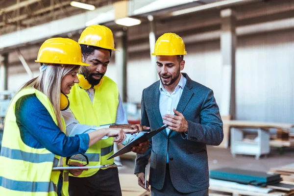 Çeşitli fabrika çalışanlarından oluşan bir ekip ve bir üretim tesisi içinde takım çalışması, liderlik ve endüstriyel güvenliği temsil eden, kask ve yüksek görünürlüklü yelek gibi koruyucu ekipmanlar giyen bir yönetici..