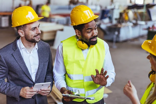 Çeşitli fabrika çalışanlarından oluşan bir ekip ve bir üretim tesisi içinde takım çalışması, liderlik ve endüstriyel güvenliği temsil eden, kask ve yüksek görünürlüklü yelek gibi koruyucu ekipmanlar giyen bir yönetici..