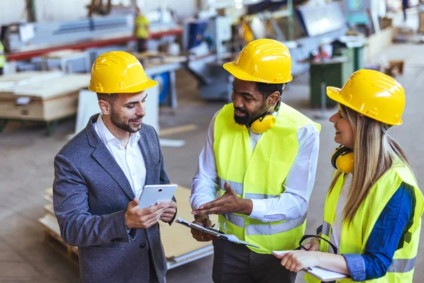 Çeşitli fabrika çalışanlarından oluşan bir ekip ve bir üretim tesisi içinde takım çalışması, liderlik ve endüstriyel güvenliği temsil eden, kask ve yüksek görünürlüklü yelek gibi koruyucu ekipmanlar giyen bir yönetici..