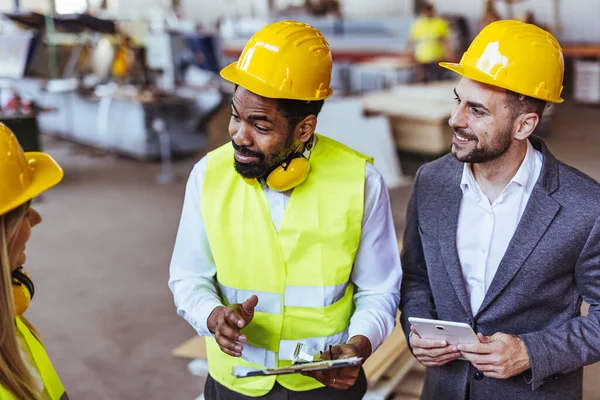 Çeşitli fabrika çalışanlarından oluşan bir ekip ve bir üretim tesisi içinde takım çalışması, liderlik ve endüstriyel güvenliği temsil eden, kask ve yüksek görünürlüklü yelek gibi koruyucu ekipmanlar giyen bir yönetici..