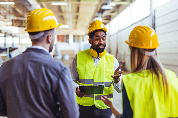Diverse team of factory employees and a manager inside a production facility, representing teamwork, leadership, and industrial safety, wearing protective equipment like helmets and high-visibility vests.