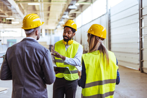 Diverse team of factory employees and a manager inside a production facility, representing teamwork, leadership, and industrial safety, wearing protective equipment like helmets and high-visibility vests.
