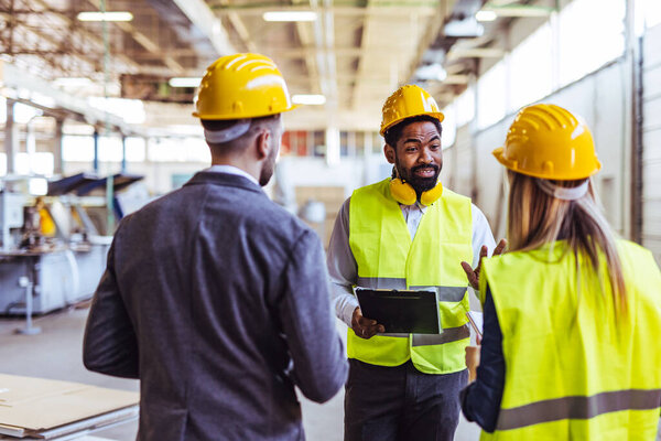 Diverse team of factory employees and a manager inside a production facility, representing teamwork, leadership, and industrial safety, wearing protective equipment like helmets and high-visibility vests.