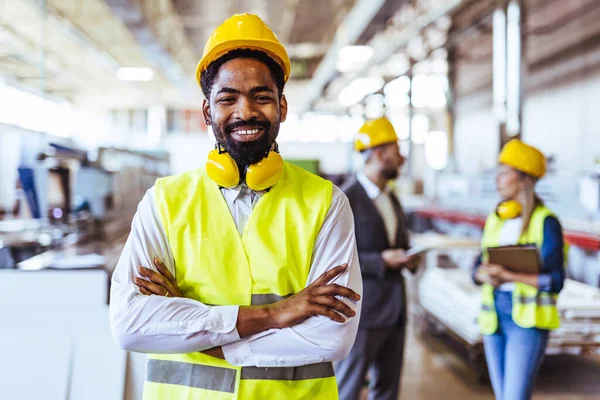 Çeşitli fabrika çalışanlarından oluşan bir ekip ve bir üretim tesisi içinde takım çalışması, liderlik ve endüstriyel güvenliği temsil eden, kask ve yüksek görünürlüklü yelek gibi koruyucu ekipmanlar giyen bir yönetici..