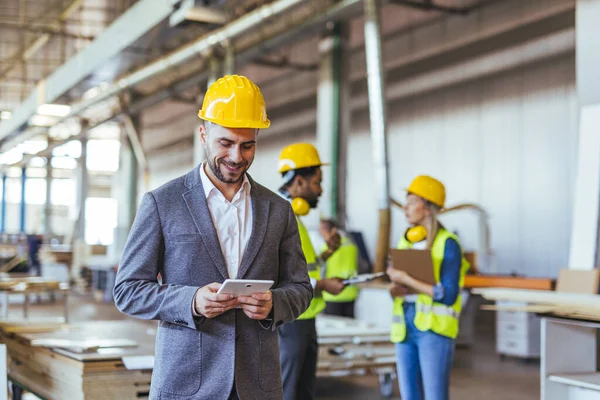 Bir fabrikada bir cihazı kontrol etmek için kasket takan bir işadamı, ekibinin yanında yüksek görünürlüklü yeleklerle ve güvenlik teçhizatıyla birlikte çalışıyor..