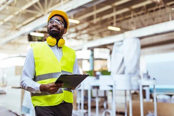 Afro-Amerikan erkek mühendis sarı kasklı, güvenlik gözlüklü ve depoda panosu olan.