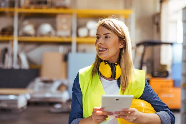 Kendine güvenen bir kadın çalışma ortamında, tablet kullanıyor, güvenlik giysileri içinde, bir depoda. Görüntü teknolojiyi, üretkenliği, modern iş etiğini ve güvenlik standartlarını vurguluyor.