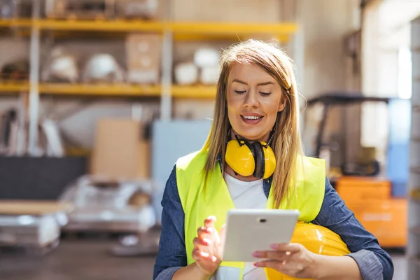 Yüksek görüş açısına sahip yelek ve kulaklıklı bir kadın işçi, inşaat ve güvenlik yönetimi teknolojisini gösteren bir depoda tablet kullanıyor..