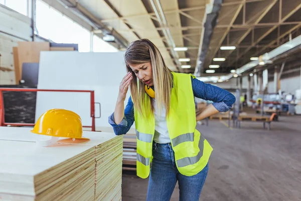Bir inşaat işçisi bir depoda yorgun görünüyor, güvenlik giysileri giyiyor. Endüstriyel iş stresi ve depo koşullarını gösteren bir kask yakındaki bir yığının üzerine yerleştiriliyor..