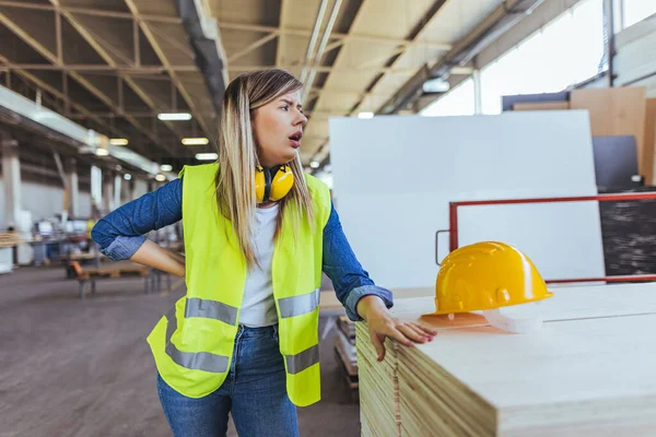 Endüstriyel bir ortamda yansıtıcı yelek ve güvenlik ekipmanı giyen genç bir kadın. Ahşap malzemeler ve sanayi araçları arka planda görünür olup, üretim veya inşaat bağlamını vurguluyor.