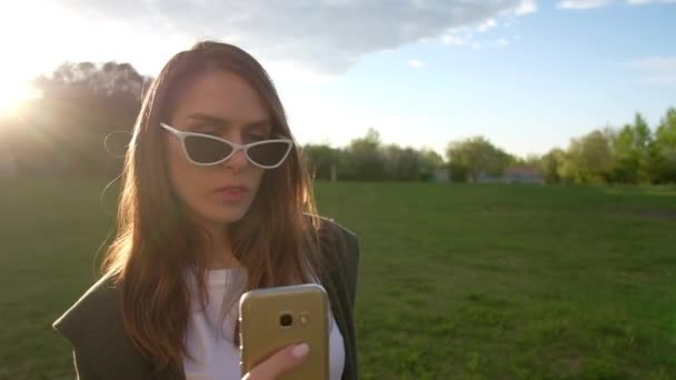 Hermosa mujer joven haciendo selfie con teléfono inteligente dorado en un parque . — Vídeos de Stock