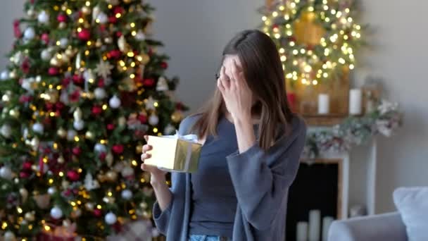 Joven mujer emocionada sosteniendo regalo, caja de sacudidas tratando de adivinar qué dentro, en el fondo del árbol de Navidad en casa decorada . — Vídeos de Stock