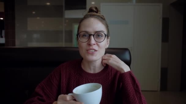 Cute young woman in eyeglasses, sitting in the cafe and talking to someone. — Stock Video