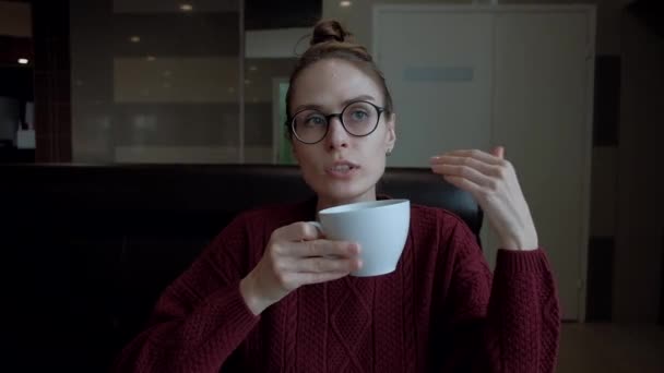 Niedliche junge Frau mit Brille, die im Café sitzt und mit jemandem spricht. — Stockvideo