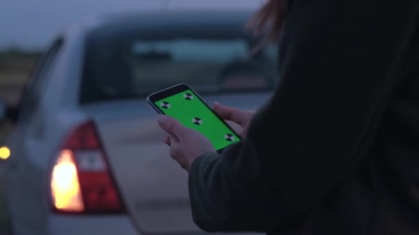 Kvinna som innehar en smartphone med grön skärm på bakgrund av trasiga bil. — Stockvideo