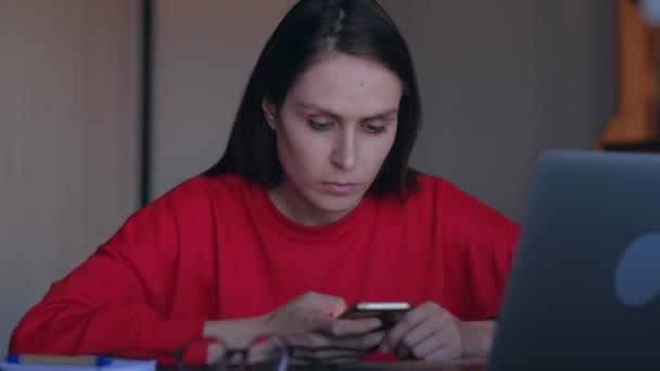 Schattige vrouw overschrijft in Smartphone informatie van de laptop. — Stockvideo