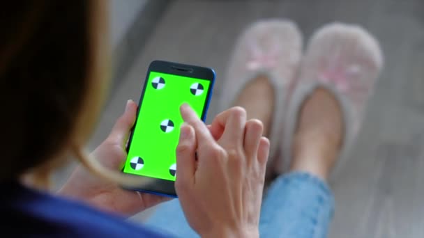 Frau in rosa Pantoffeln mit Smartphone mit grünem Bildschirm auf dem Boden sitzend. — Stockvideo
