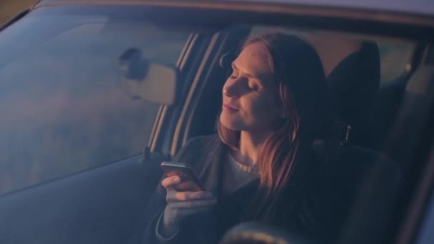 Mujer sentarse en el coche utiliza un teléfono inteligente con ganas de disfrutar de la puesta del sol . — Vídeos de Stock