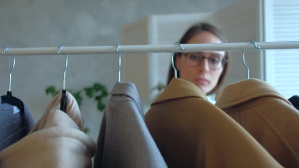 Young woman in glasses chooses clothes that hanging on a clothing rack in a shop — Stock Video