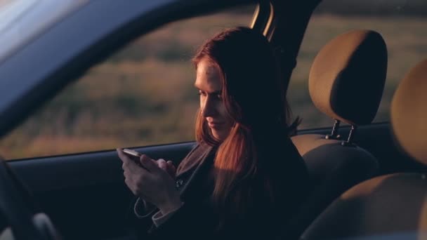 Mujer sentarse en el coche utiliza un teléfono inteligente en la noche antes de la puesta del sol . — Vídeos de Stock
